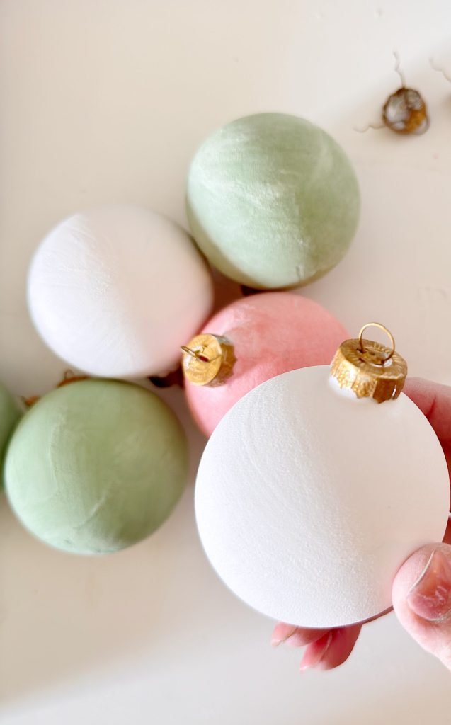 A hand holds a white Christmas ornament next to several pastel-colored ornaments in white, green, and pink on a light surface.