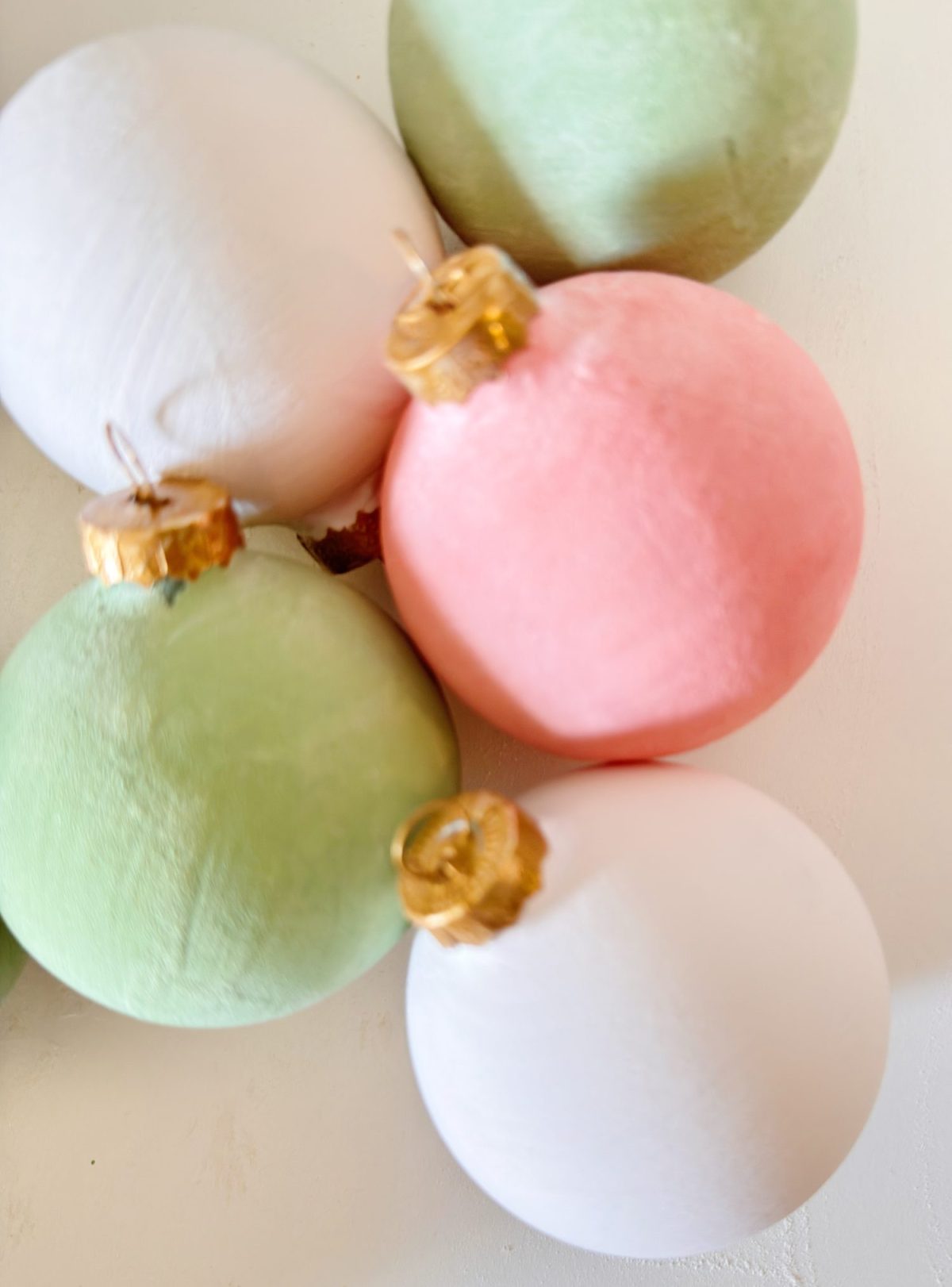 Five Christmas ornaments in pastel colors—pink, green, and white—clustered together on a white surface, each with a gold hanging cap.