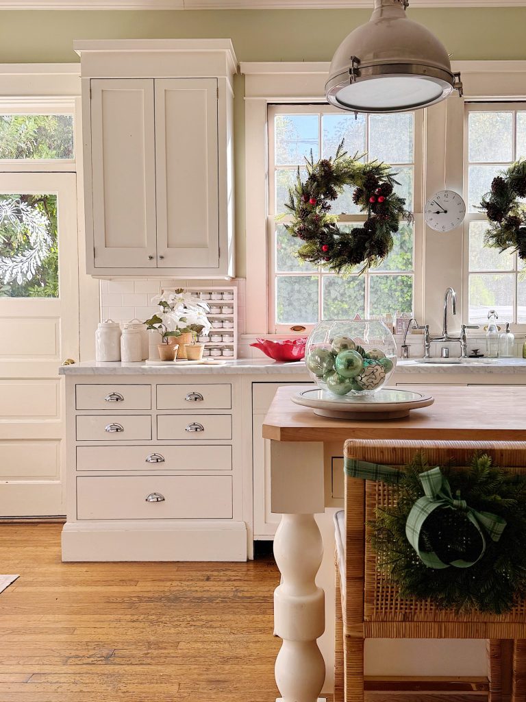 Bright kitchen with white cabinets and wood floor, decorated with holiday wreaths, a glass bowl of ornaments, and a bowl of apples on the counter. Sunlight streams through the windows.