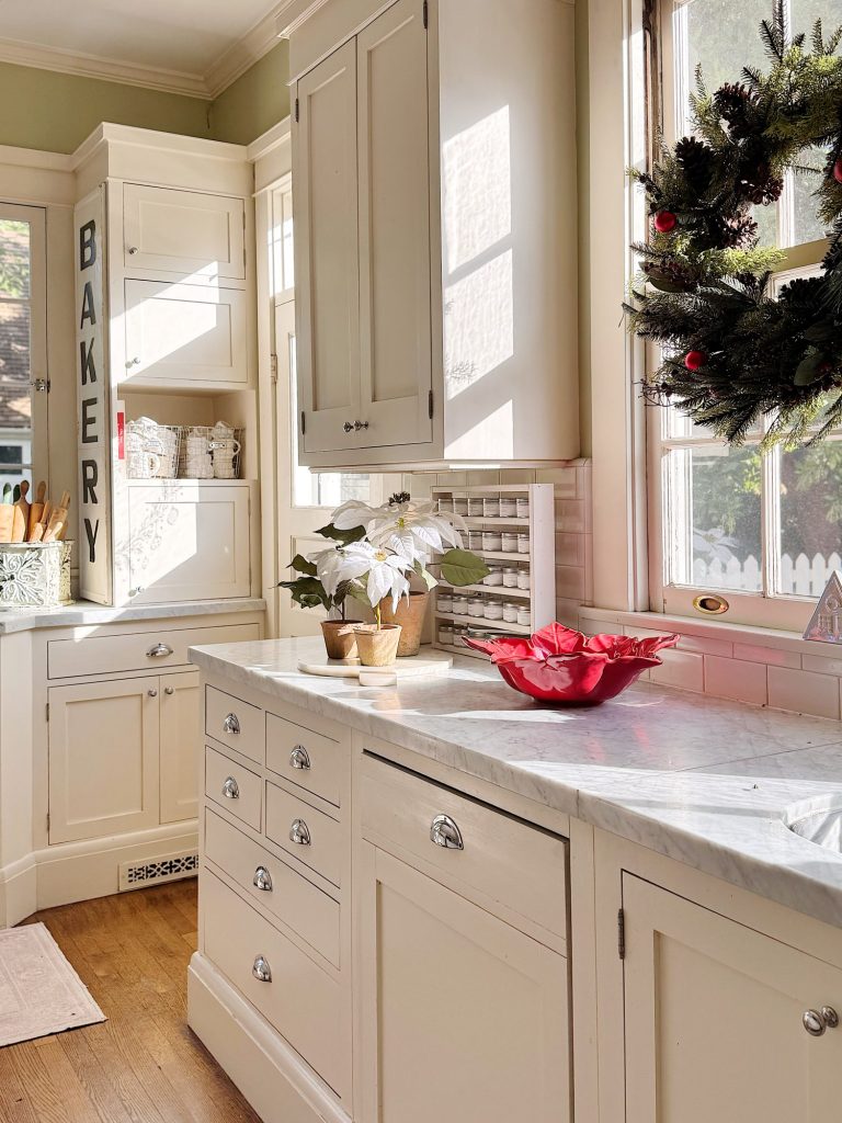 Bright kitchen with white cabinets, marble countertops, a red dish, white poinsettia plant, "BAKERY" sign, and a decorated holiday wreath by the window.