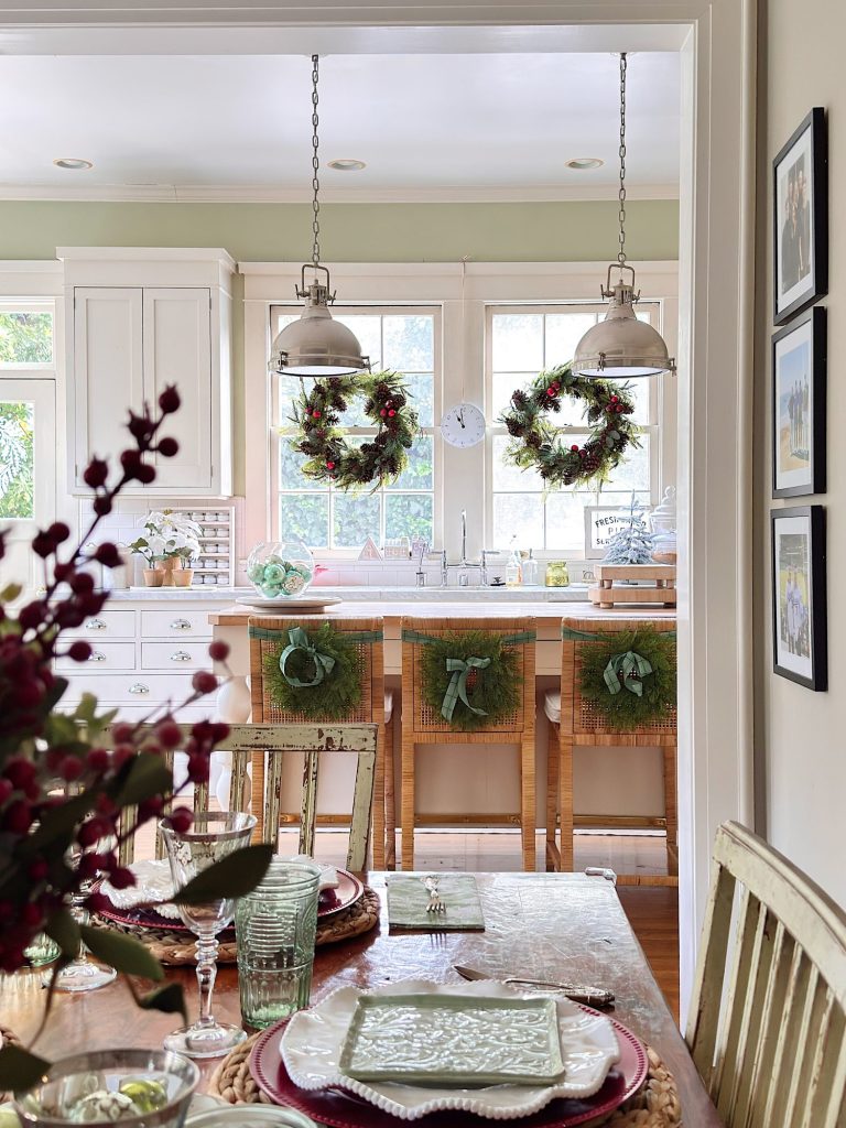 A kitchen decorated for the holidays with wreaths hanging above the island, on barstools, and on the window; a dining table is set in the foreground.