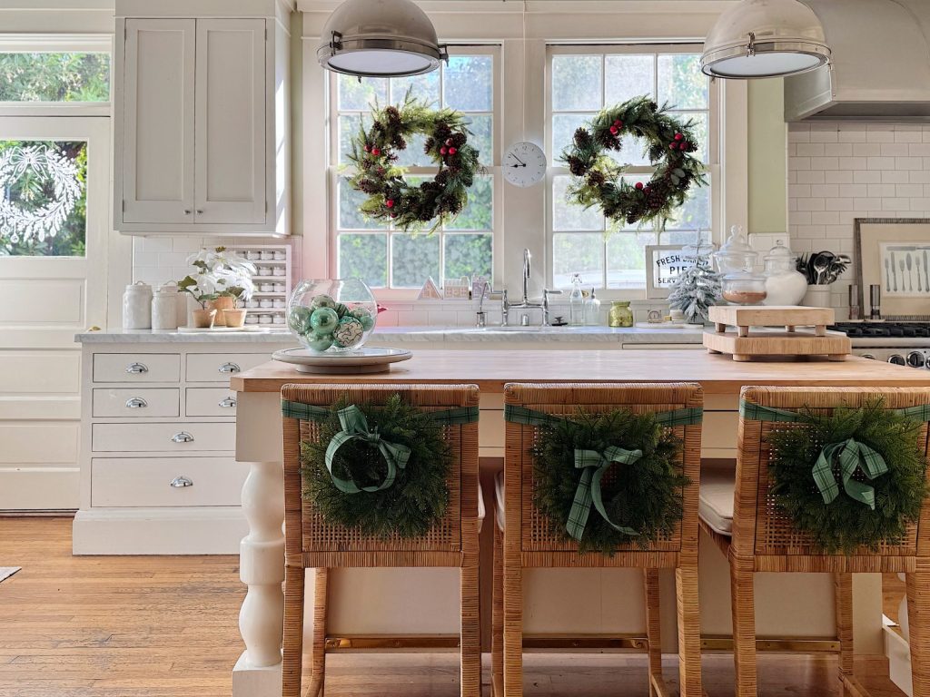 A bright kitchen with wreaths on the window and chairs, white cabinets, wood floors, and a central island with glass jars and a bowl of ornaments.