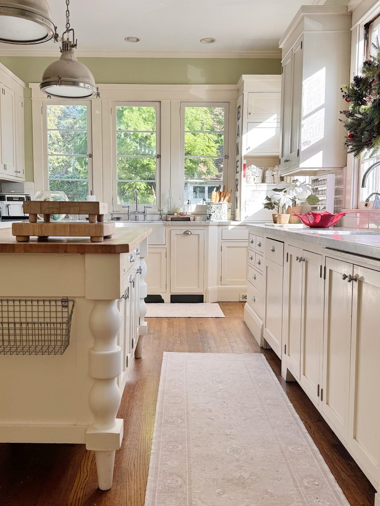 Bright kitchen with white cabinets, wooden floor, central island, large windows, and sunlight streaming in; various kitchen items and a small plant are visible on the counters.