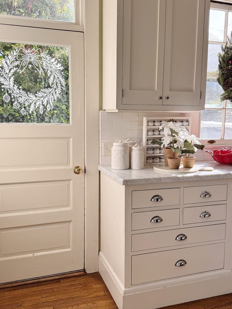A bright kitchen corner with white cabinets, a marble countertop, potted plants, ceramic canisters, and a windowed door decorated with a white holiday wreath design.