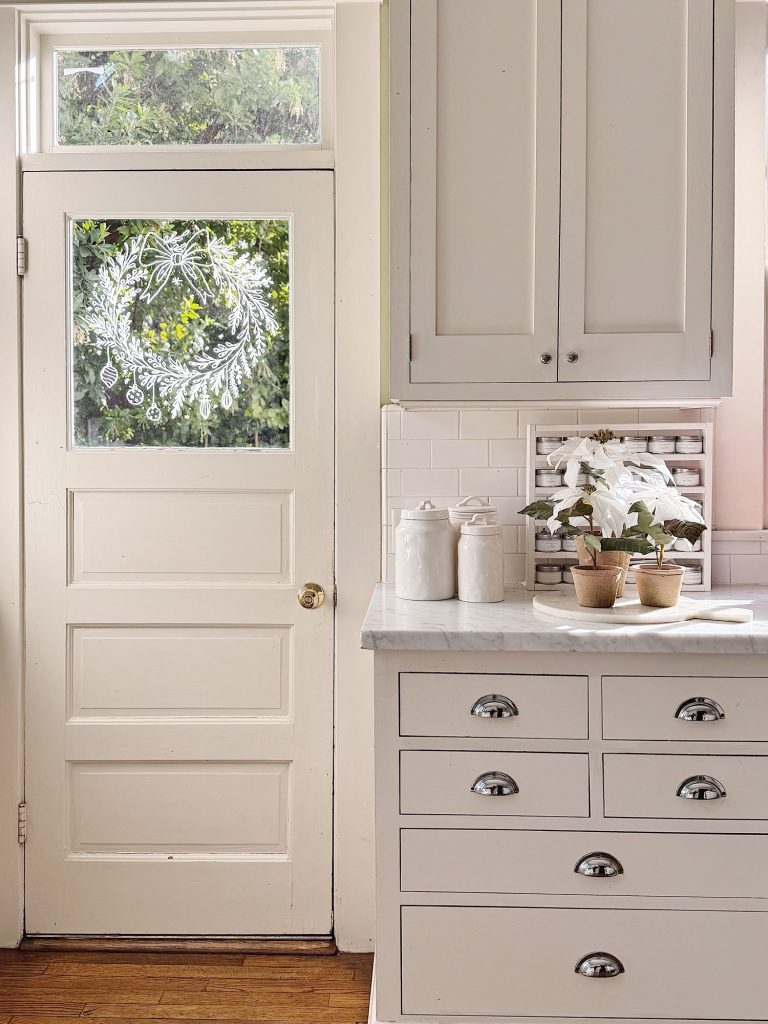 A white kitchen with marble countertops, potted plants, and canisters; a back door with a frosted holiday wreath design is visible.