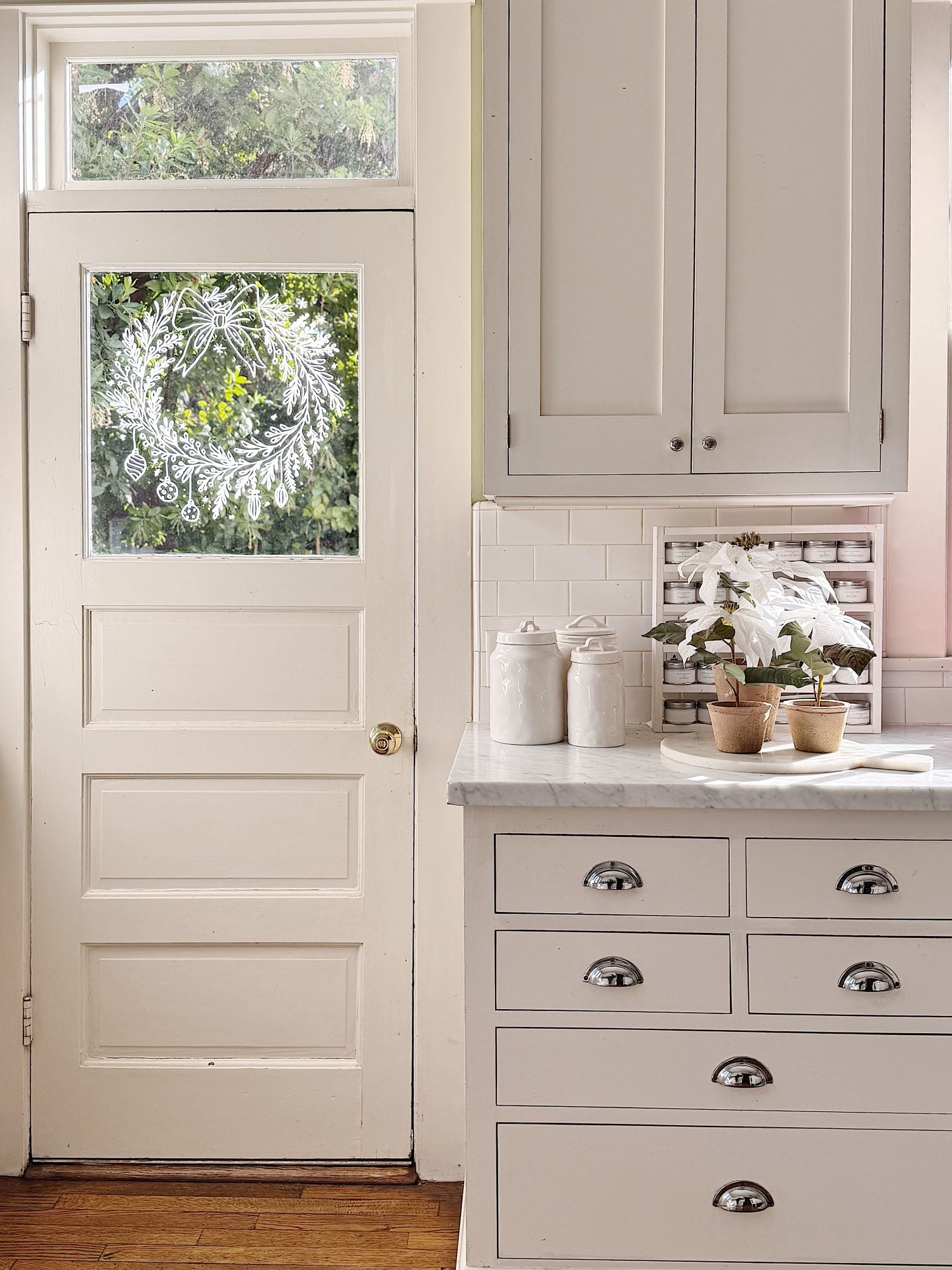 A white kitchen with marble countertops, potted plants, and canisters; a back door with a frosted holiday wreath design is visible.
