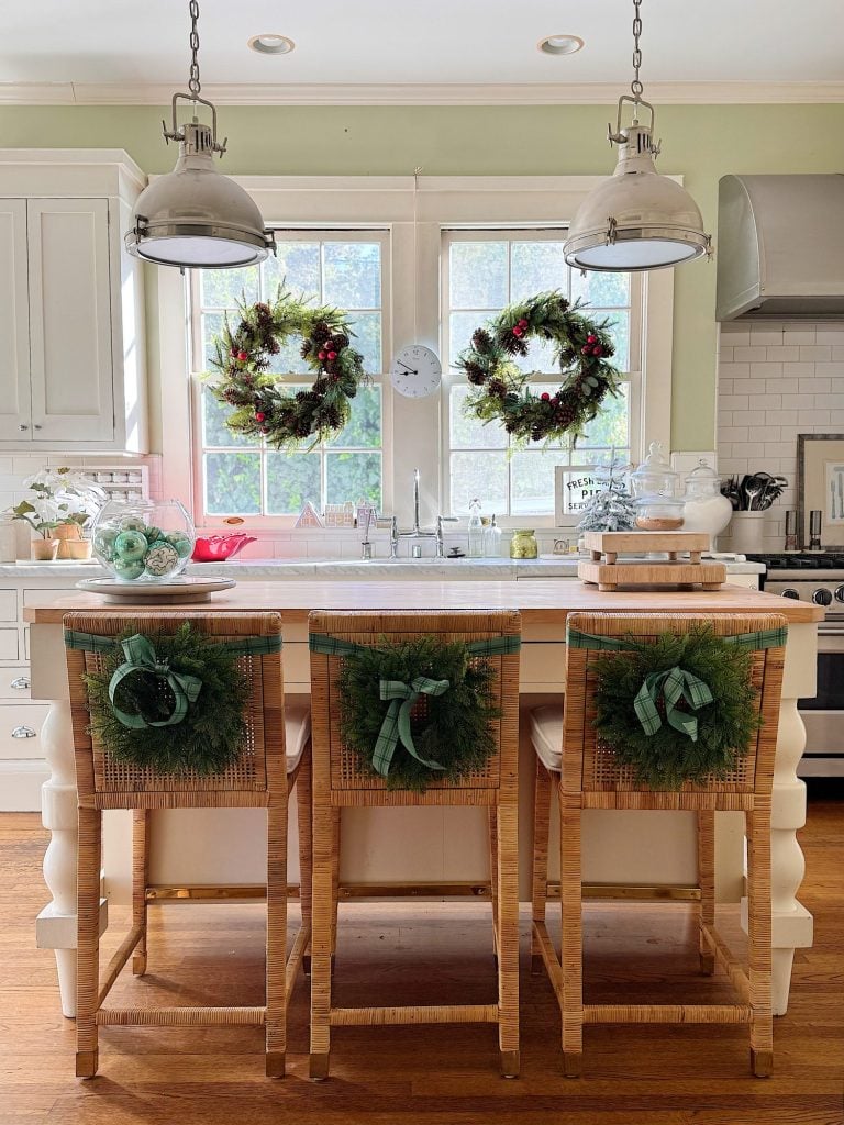 A bright kitchen decorated for the holidays with wreaths on the windows and backs of three barstools, white cabinets, and pendant lights above a central island.