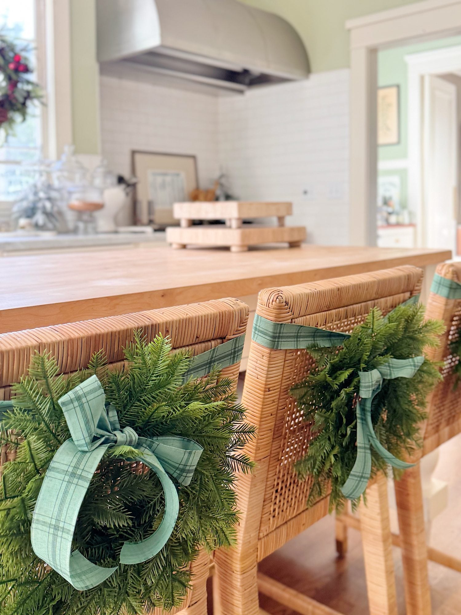 Wicker chairs at a wooden kitchen island are decorated with small green wreaths and plaid ribbons. White kitchen cabinets and a range hood are visible in the background.