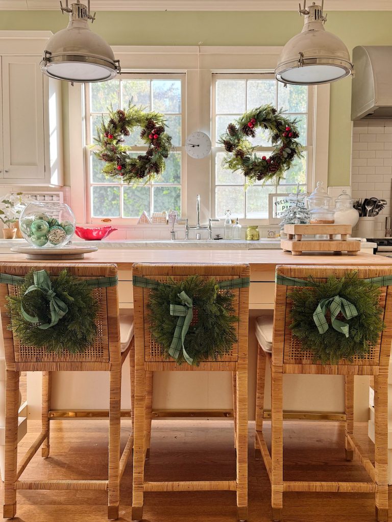 Three wicker barstools with green wreaths and ribbons are lined up at a kitchen island; two windows above the sink also display holiday wreaths.