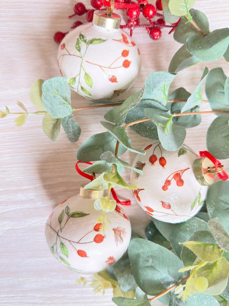 Three white Christmas ornaments with red and green botanical designs hang among green eucalyptus leaves and red berries against a light wooden background.