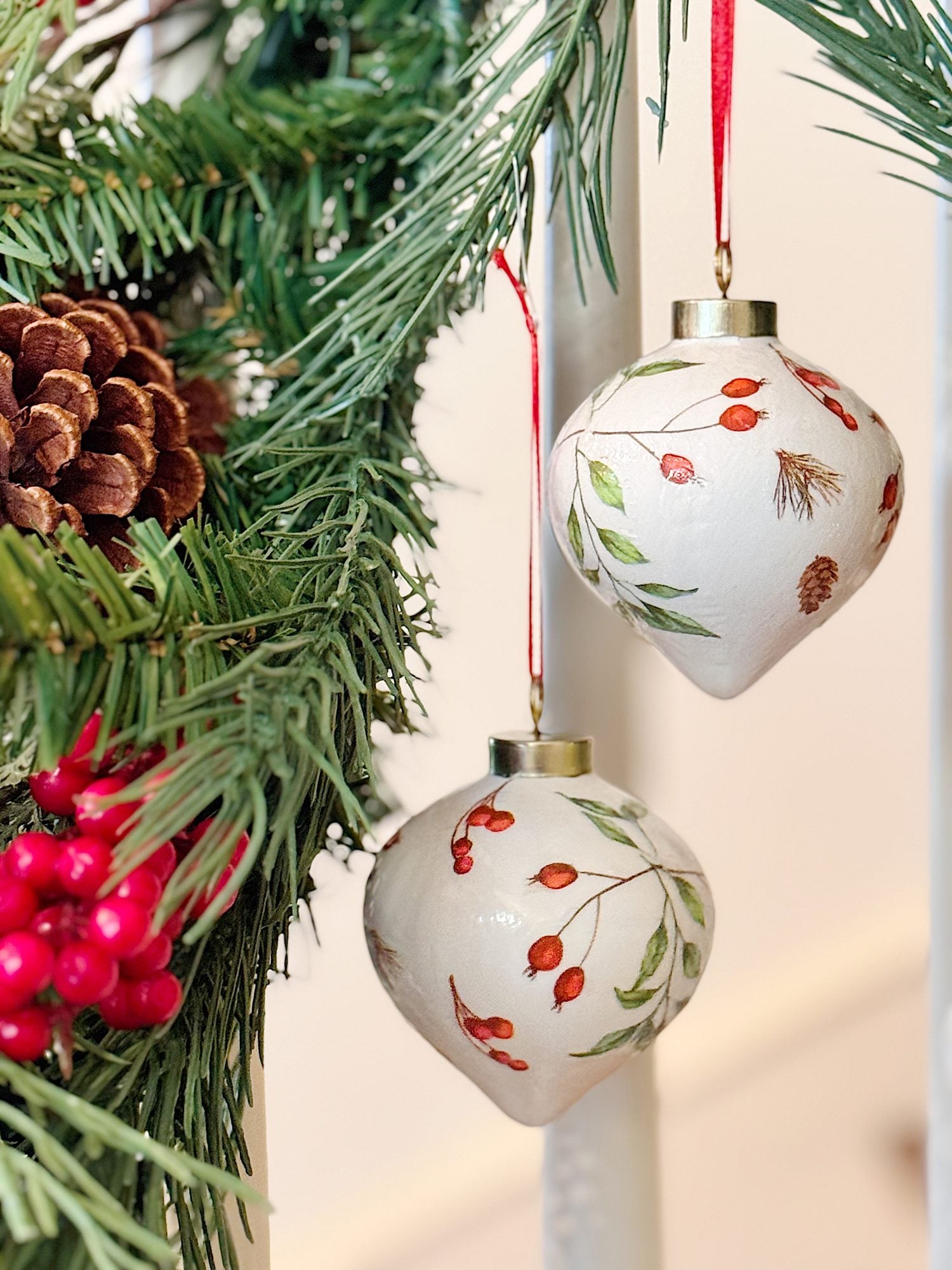 Two white Christmas ornaments with botanical designs hang from red ribbons on a pine garland with pinecones and red berries.