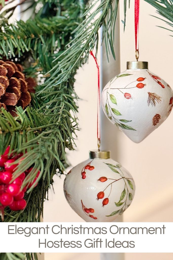 Two white Christmas ornaments with botanical designs hang on a pine branch with pinecones and red berries; text reads "Elegant Christmas Ornament Hostess Gift Ideas.