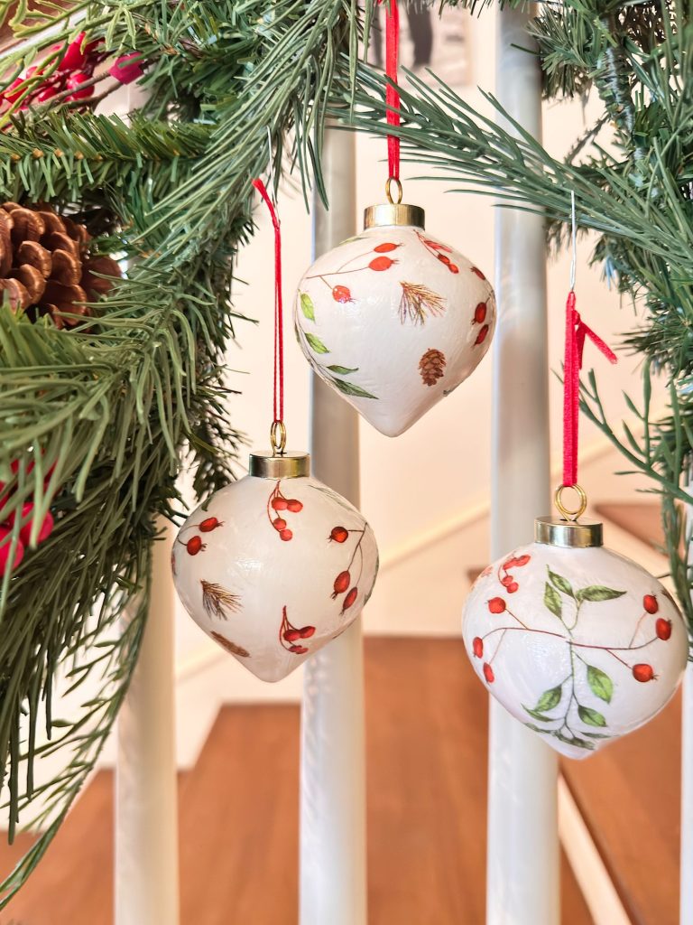 Three white Christmas ornaments with red and green botanical designs hang from a pine garland on a staircase railing.