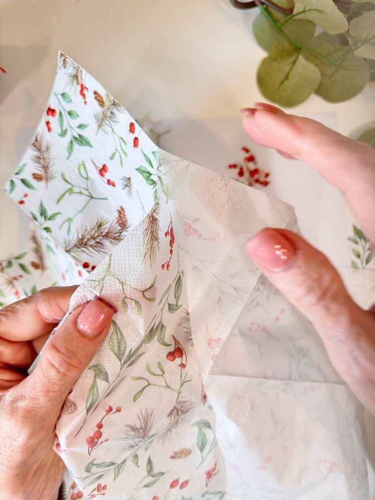 Two hands separate layers of a decorative paper napkin with a floral and berry pattern on a white surface.