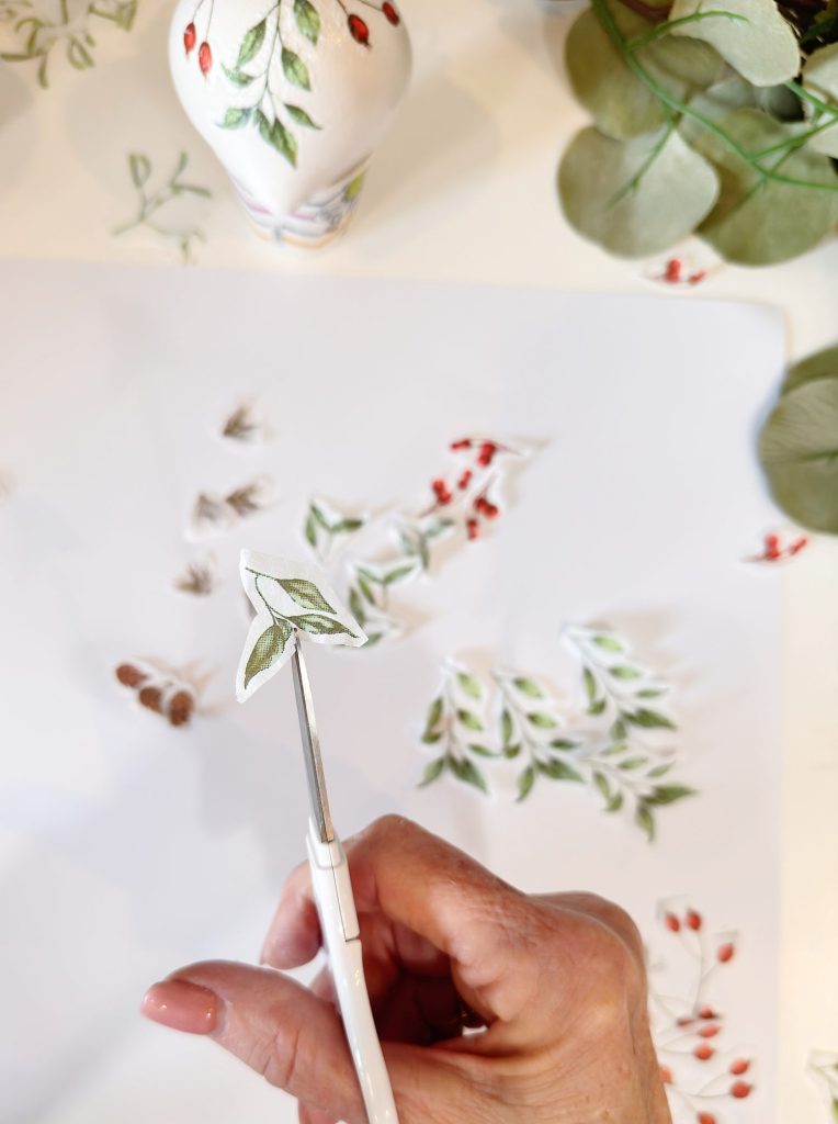 A hand uses scissors to cut out a paper illustration of green leaves, with more botanical drawings and cutouts on a white surface in the background.