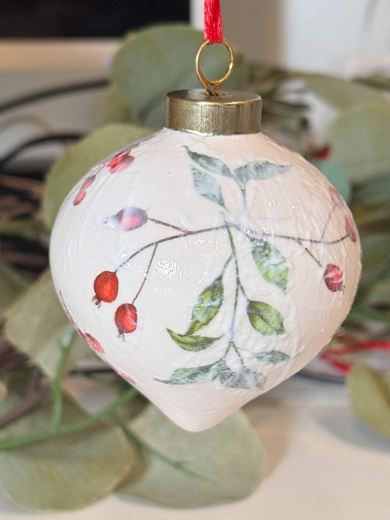 A white Christmas ornament with a botanical design featuring green leaves and red berries hangs from a red ribbon.