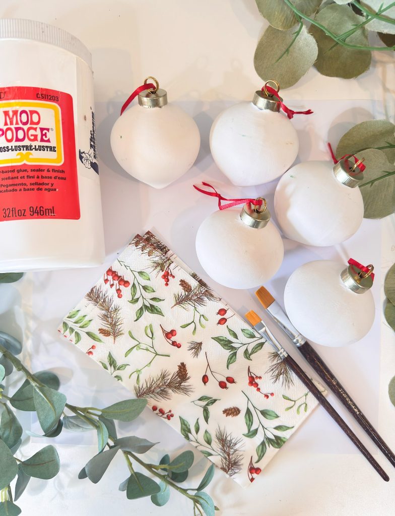 A bottle of Mod Podge, five plain white ornaments with red strings, a decorative napkin, two brushes, and green foliage on a white surface.