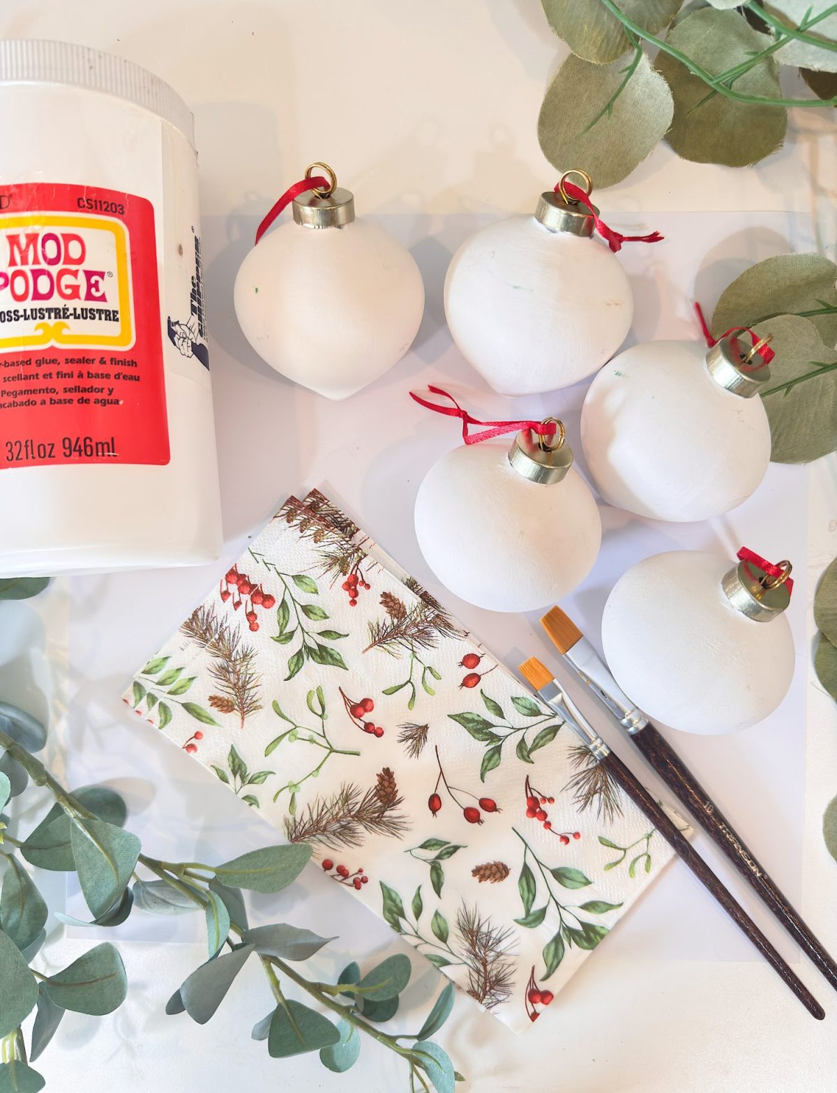 A bottle of Mod Podge, five plain white ornaments with red strings, a decorative napkin, two brushes, and green foliage on a white surface.