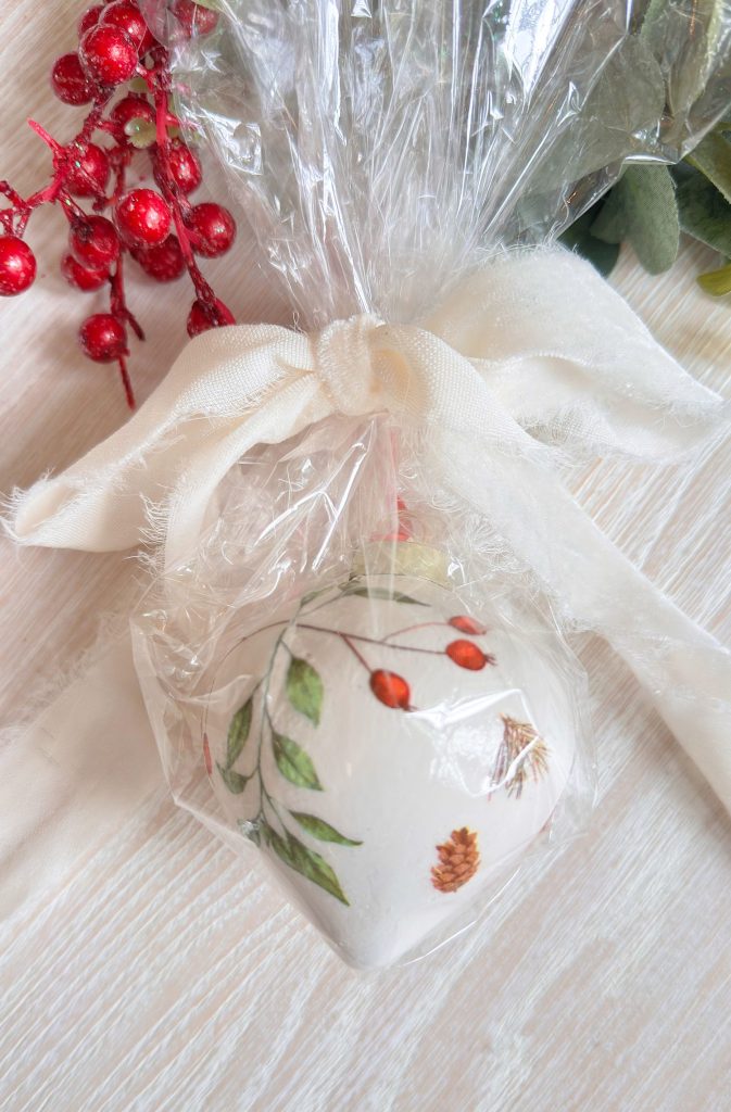 A white ornament with painted leaves, berries, and pinecones is wrapped in clear plastic tied with a white ribbon, next to red decorative berries on a light wood surface.