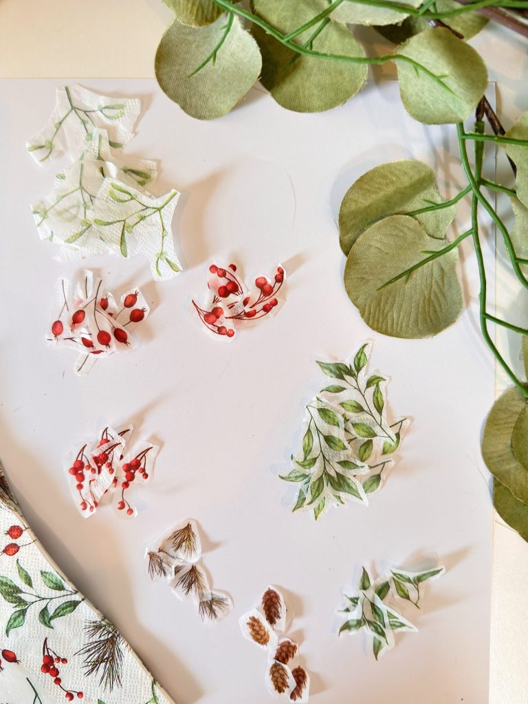A white surface with cut-out illustrations of leaves, pinecones, and red berries, with green artificial leaves in the top right corner.