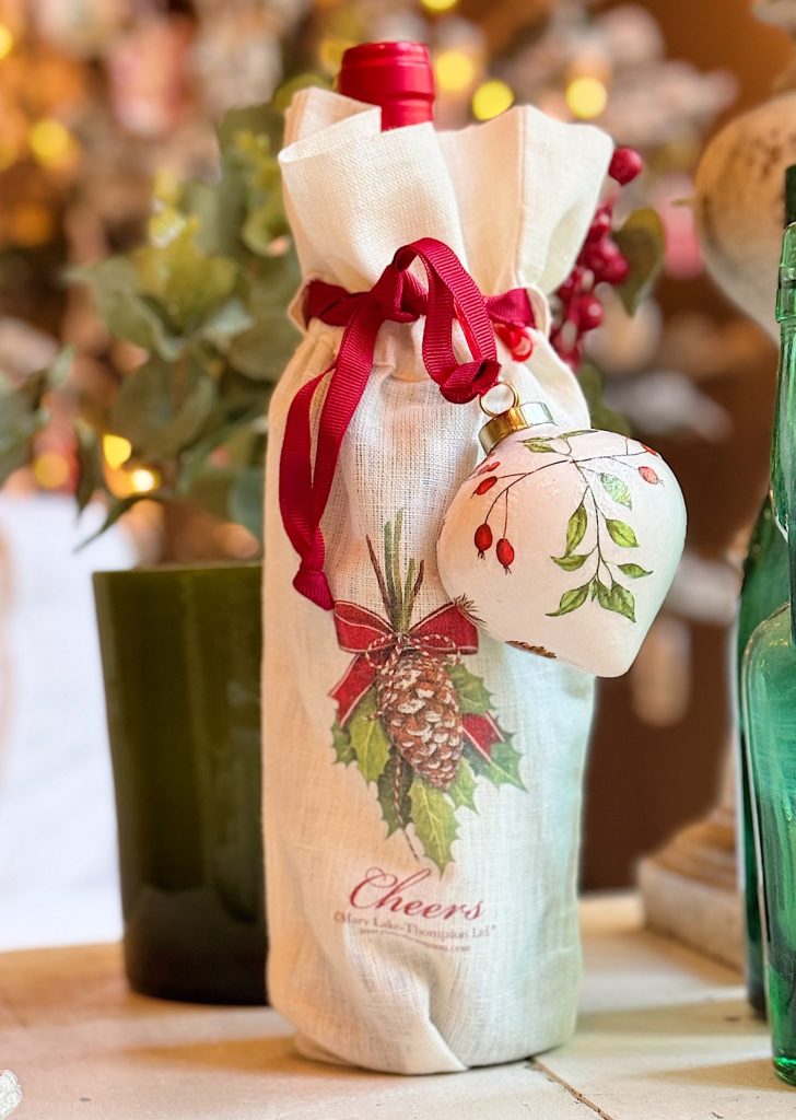 A wine bottle wrapped in a holiday-themed cloth bag with red ribbon and a decorative heart ornament, set on a table with a green glass bottle and leafy background.