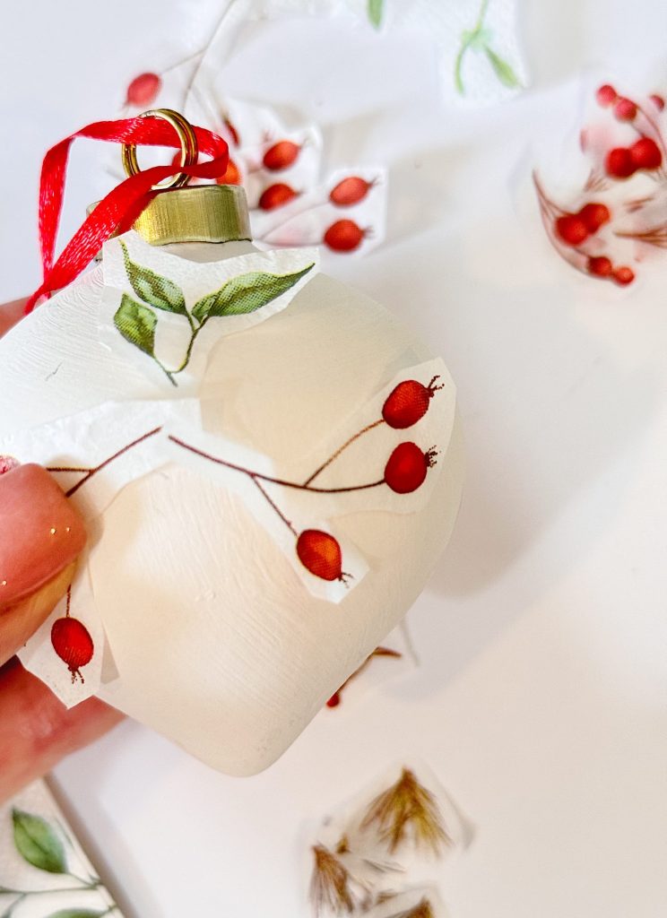 A hand holds a white ornament being decorated with cut-out botanical illustrations of red berries and green leaves, with a red ribbon attached.