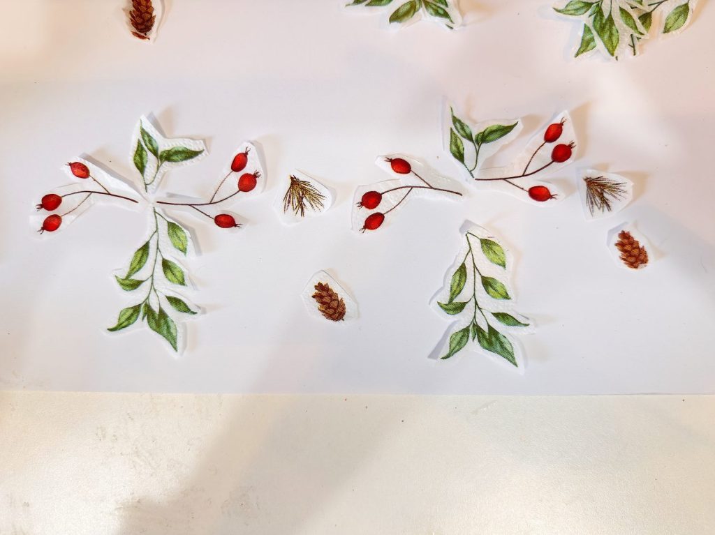 Cut-out illustrations of green leaves, red berries, pine needles, and pine cones are arranged on a white surface.