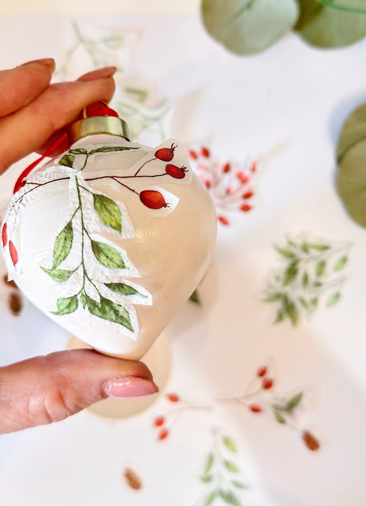 A hand holds a white Christmas ornament painted with green leaves and red berries, matching botanical artwork on the paper underneath.