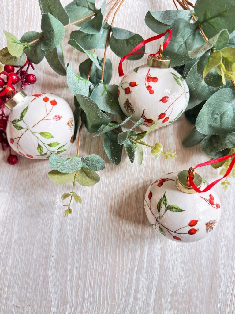 Three white Christmas ornaments with red berry designs and red ribbons are displayed on a light wood surface with green foliage and red berries.