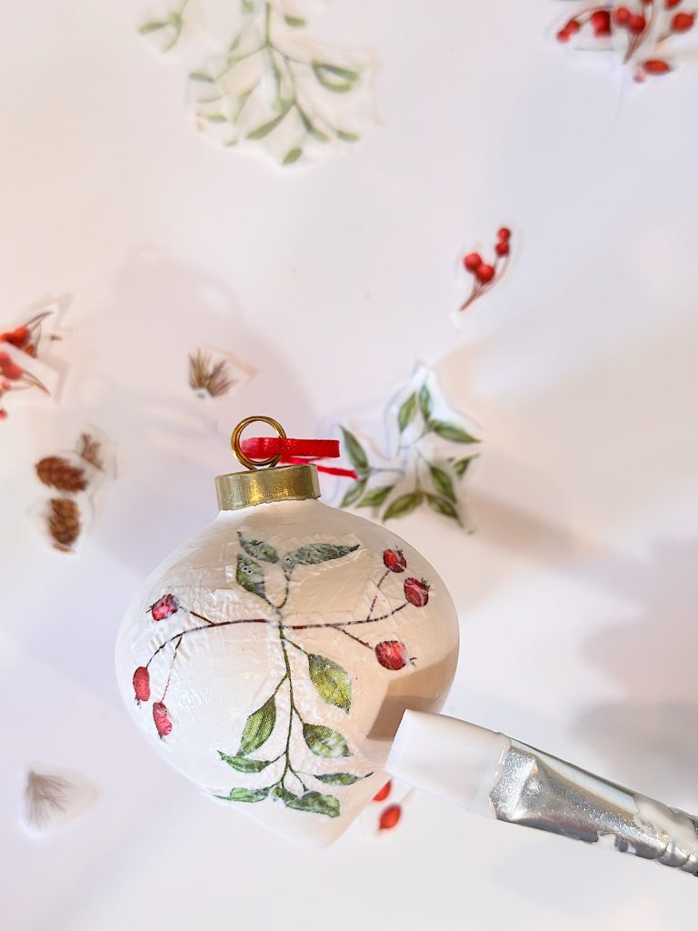 A white Christmas ornament with painted red berries and green leaves is held by a clamp, surrounded by small botanical stickers on a white surface.