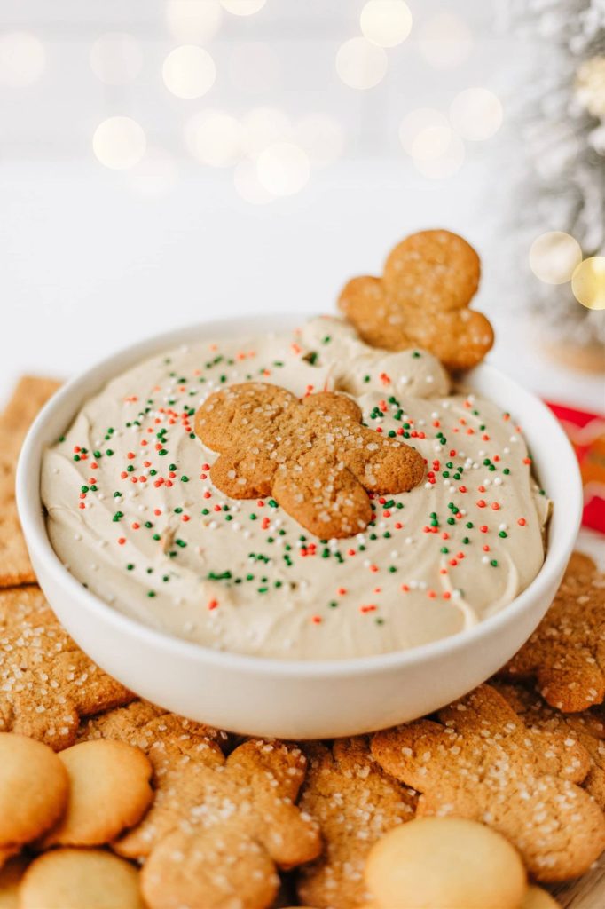 A bowl of creamy dip topped with colorful sprinkles and gingerbread cookies, surrounded by assorted cookies on a table.