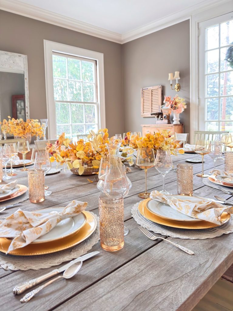 A dining table set for a meal with yellow floral centerpieces, patterned napkins, gold-rimmed plates, Mercury Glass Ornaments, glassware, and natural light coming through large windows.