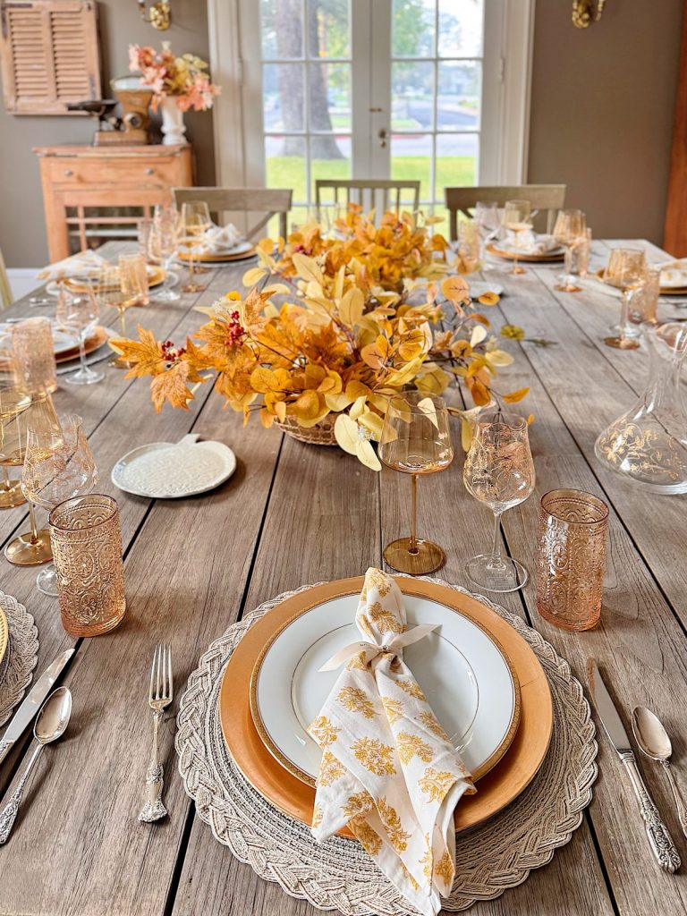 A wooden dining table is set with gold and white plates, patterned napkins, gold flatware, yellow floral centerpieces, and sparkling mercury glass ornaments, with glasses and decorative details arranged neatly.