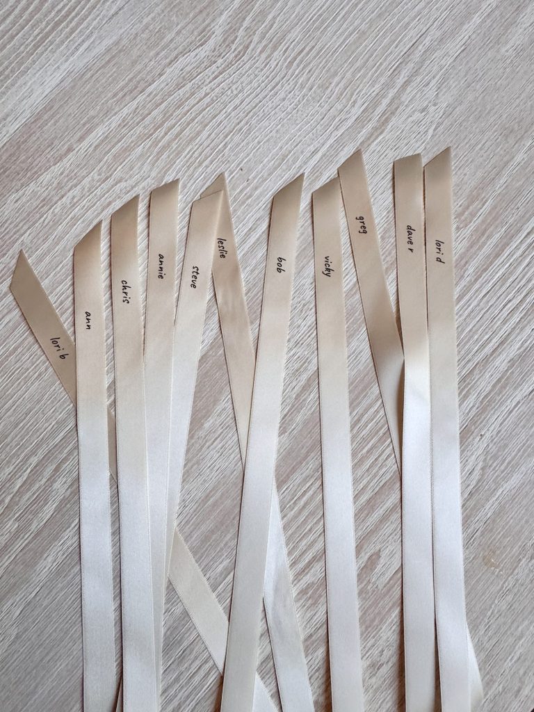 Nine cream-colored satin ribbon samples labeled with different shades are laid out on a light wood surface. Each ribbon has a name written on it in black ink.