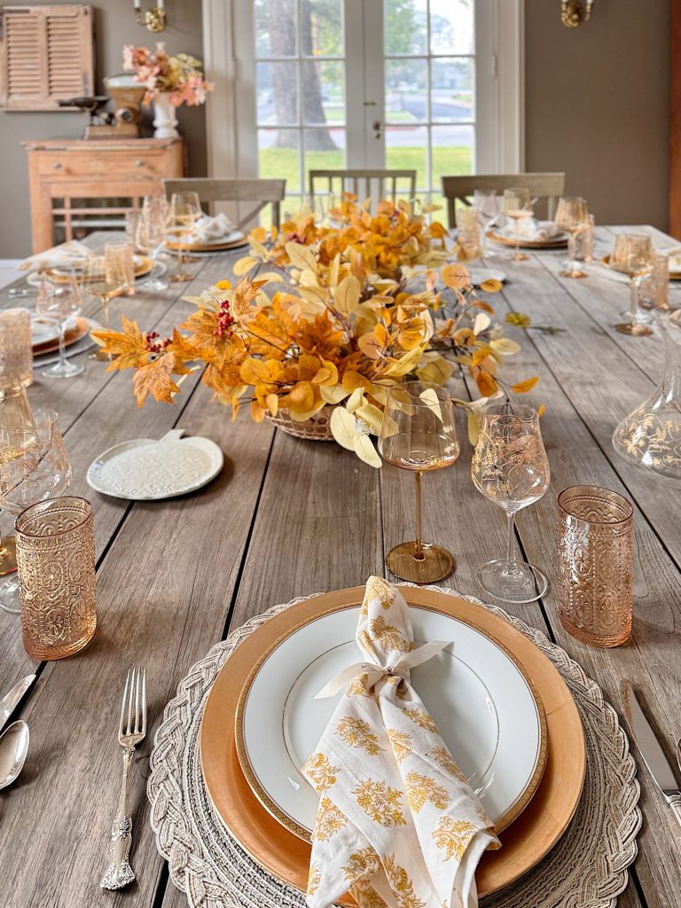 A wooden dining table set for a meal with gold-rimmed plates, patterned napkins, glassware, and a centerpiece of autumn leaves.