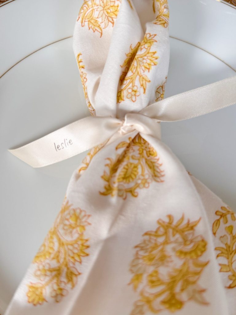 A white plate with a patterned yellow and white napkin tied with a cream ribbon, featuring the name "leslie" printed on it.