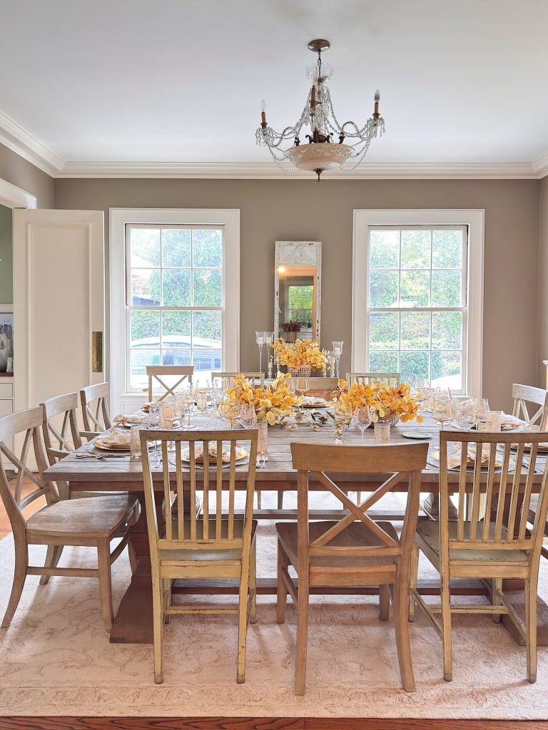 A dining room with a wooden table set for a meal, surrounded by mismatched chairs, yellow floral centerpieces, large windows, and a chandelier overhead.