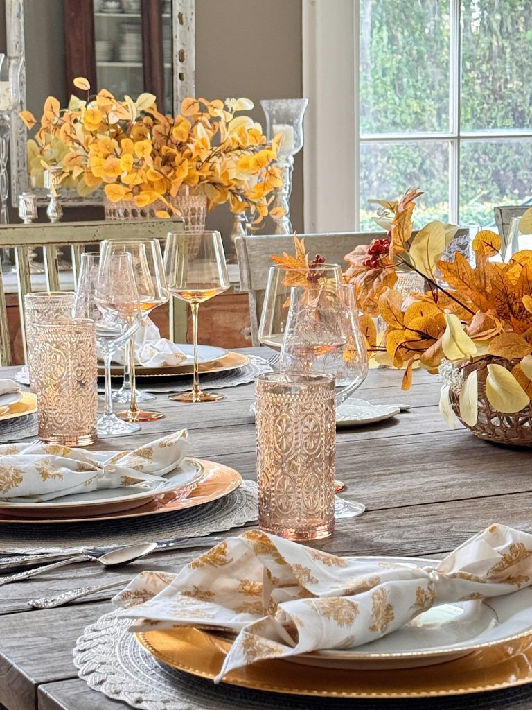 A wooden dining table set with gold-rimmed plates, patterned napkins, clear glasses, wine glasses, and fall-themed floral centerpieces.