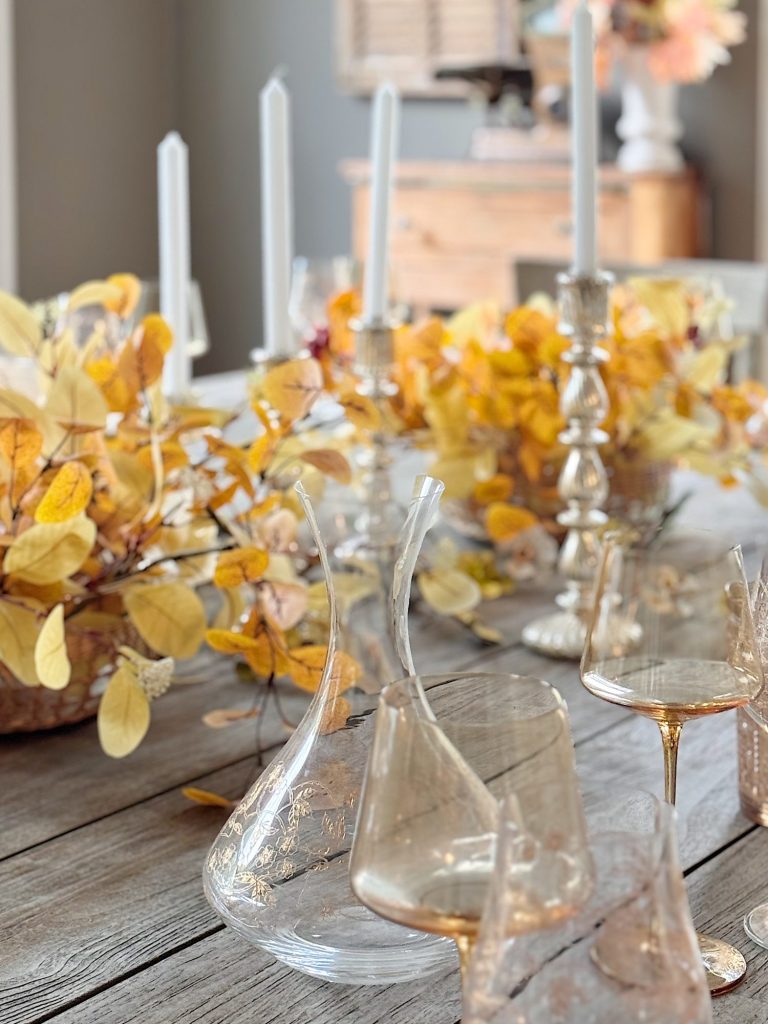 A rustic wooden table set with glassware, decanters, tall white candles, and autumn-themed floral centerpieces with yellow and orange leaves.