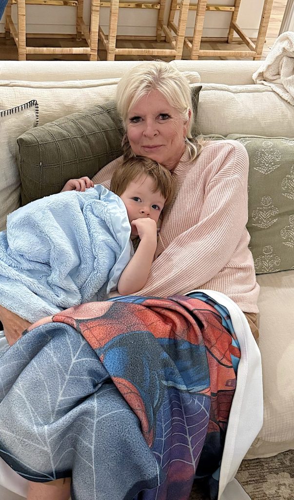 An older woman sits on a couch holding a young child wrapped in a blue blanket, both appearing relaxed and cozy under a patterned blanket, surrounded by festive Christmas decor.