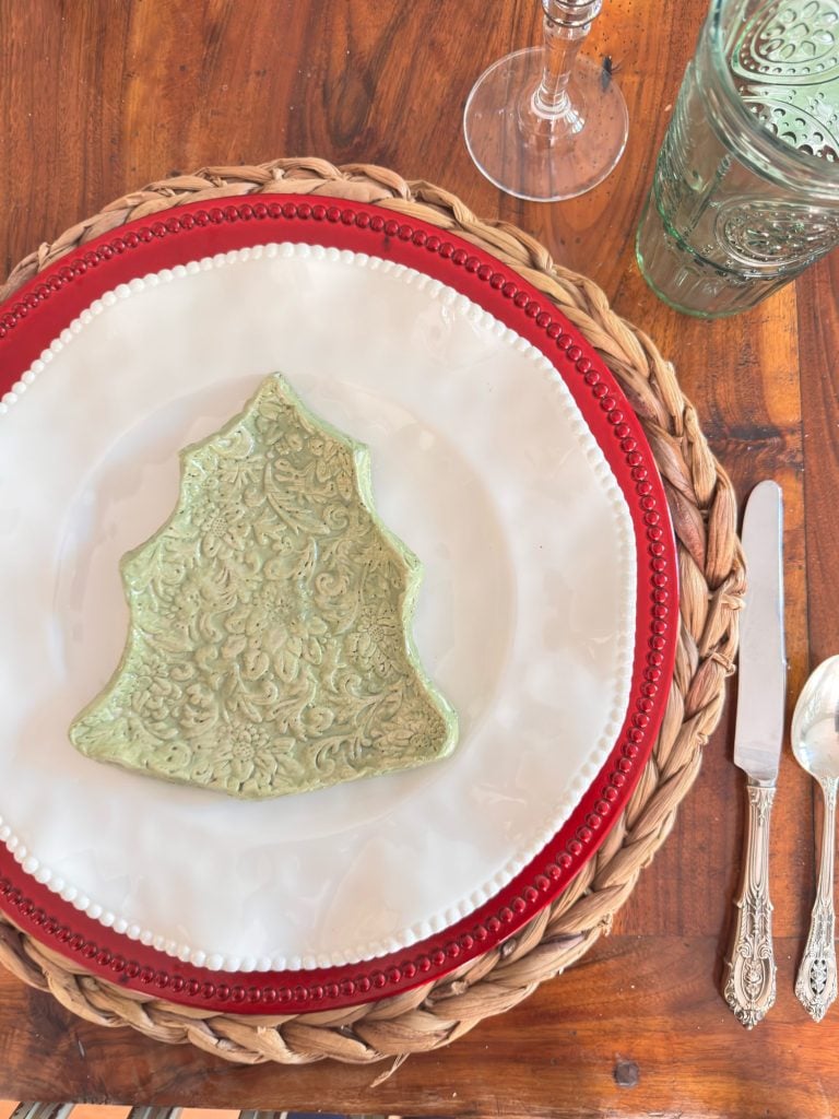 A table setting with a woven placemat, red and white plates, a green tree-shaped plate, cutlery, a green glass, and a wine glass on a wooden surface.