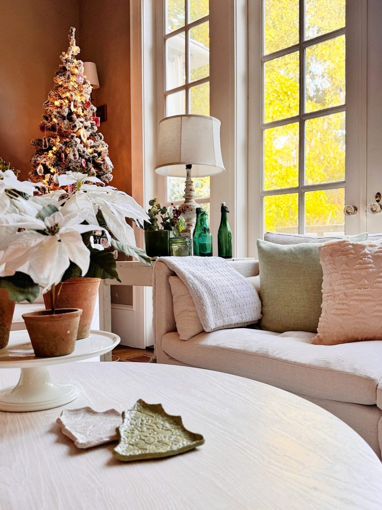 A living room with a white sofa, green and white cushions, potted white poinsettias, a decorated Christmas tree, and large windows letting in bright natural light.