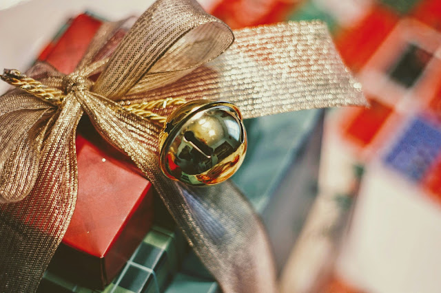 A stacked gift box wrapped in ribbon with a metallic gold bell ornament, set against a blurred background of colorful patterns.
