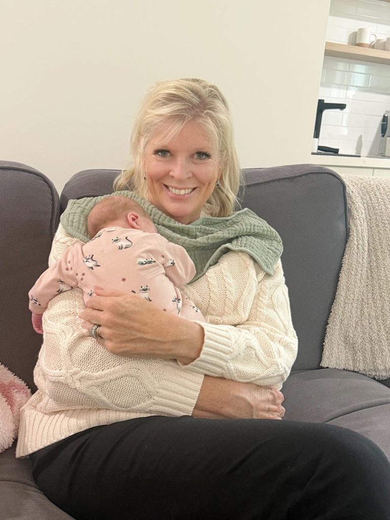 A woman sits on a couch smiling, holding a sleeping baby dressed in a pink floral outfit, as shimmering Mercury Glass Ornaments glisten softly in the background.