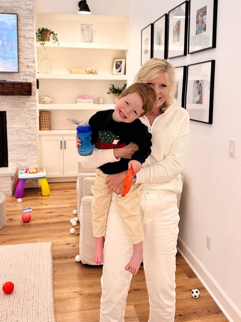 An older woman in white holds a smiling young boy with a blue sippy cup in a living room decorated with toys, framed photos, and shimmering Mercury Glass Ornaments.