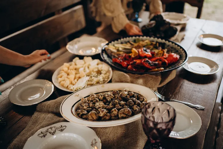 A rustic wooden table set with plates of grilled vegetables, mushrooms, cheese, and empty dishes, with hands reaching for food.