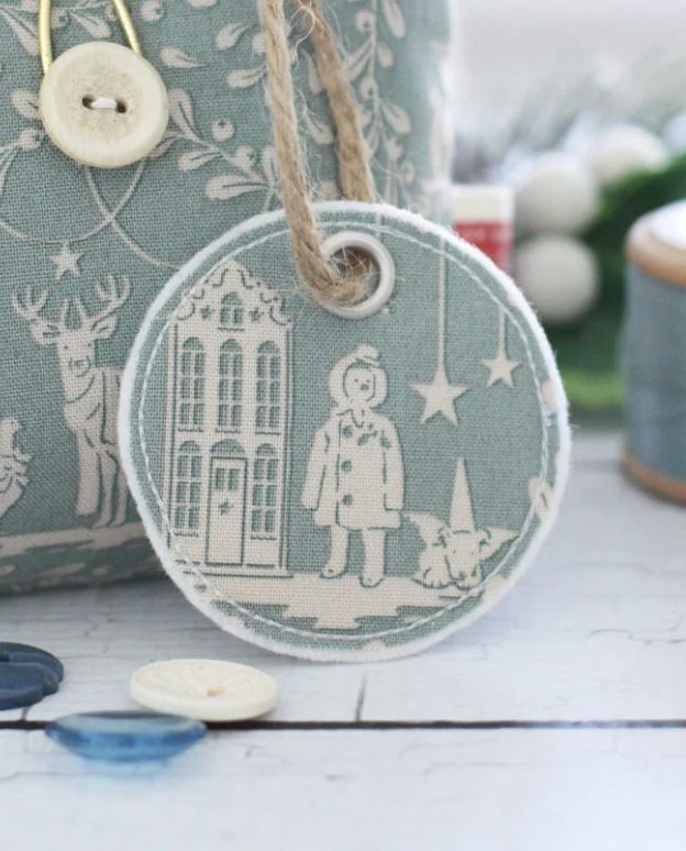 A round fabric tag with a winter scene, including a building, a person in a coat, a small dog, and stars, hanging from a twine loop. A button and spool of thread are nearby.