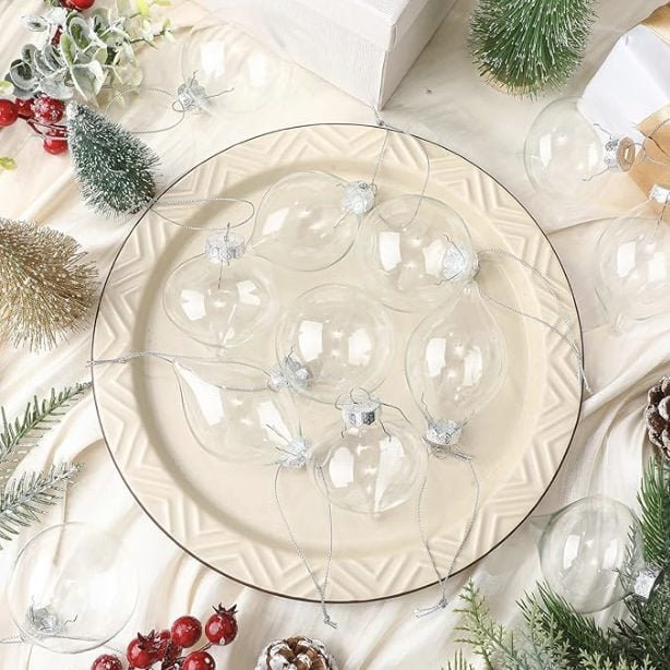 A plate with clear glass Christmas ornaments arranged on it, surrounded by artificial pine branches, berries, and small decorated trees.