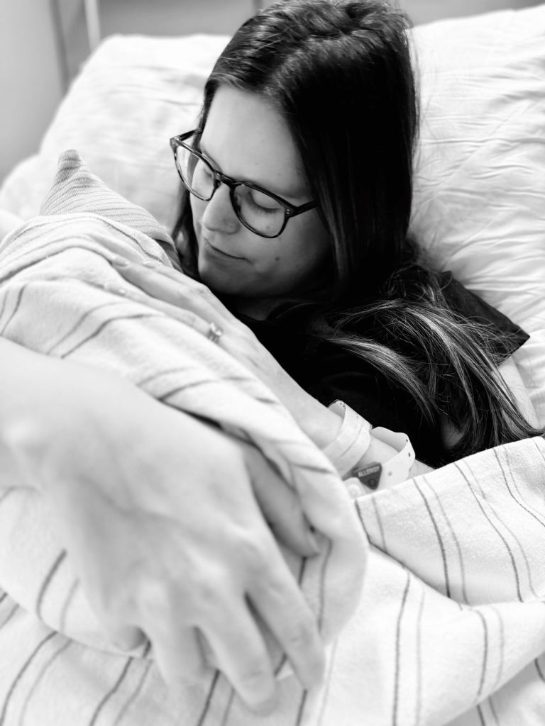 A woman with glasses lies in a hospital bed, holding a swaddled newborn in her arms, while mercury glass ornaments shimmer softly on the windowsill nearby.