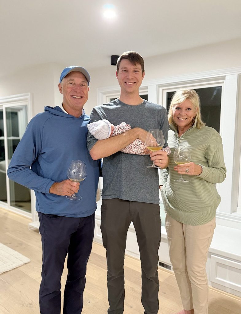 Three adults stand indoors smiling at the camera, each holding a wine glass beneath sparkling Mercury Glass Ornaments. The person in the center is holding a swaddled baby.